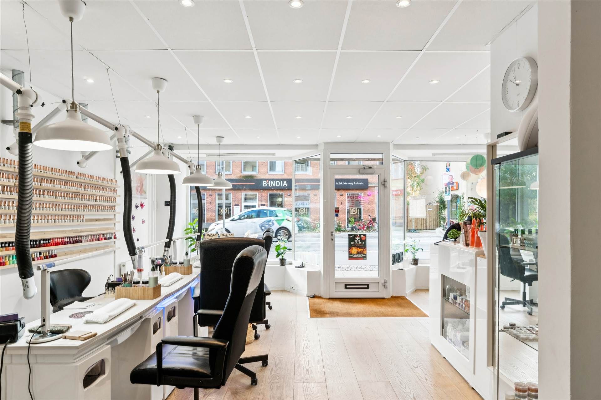 En salon med mange stole og stole med en bord og hylder med neglelak. Væggene er hvide og der er lysekroner hængende fra