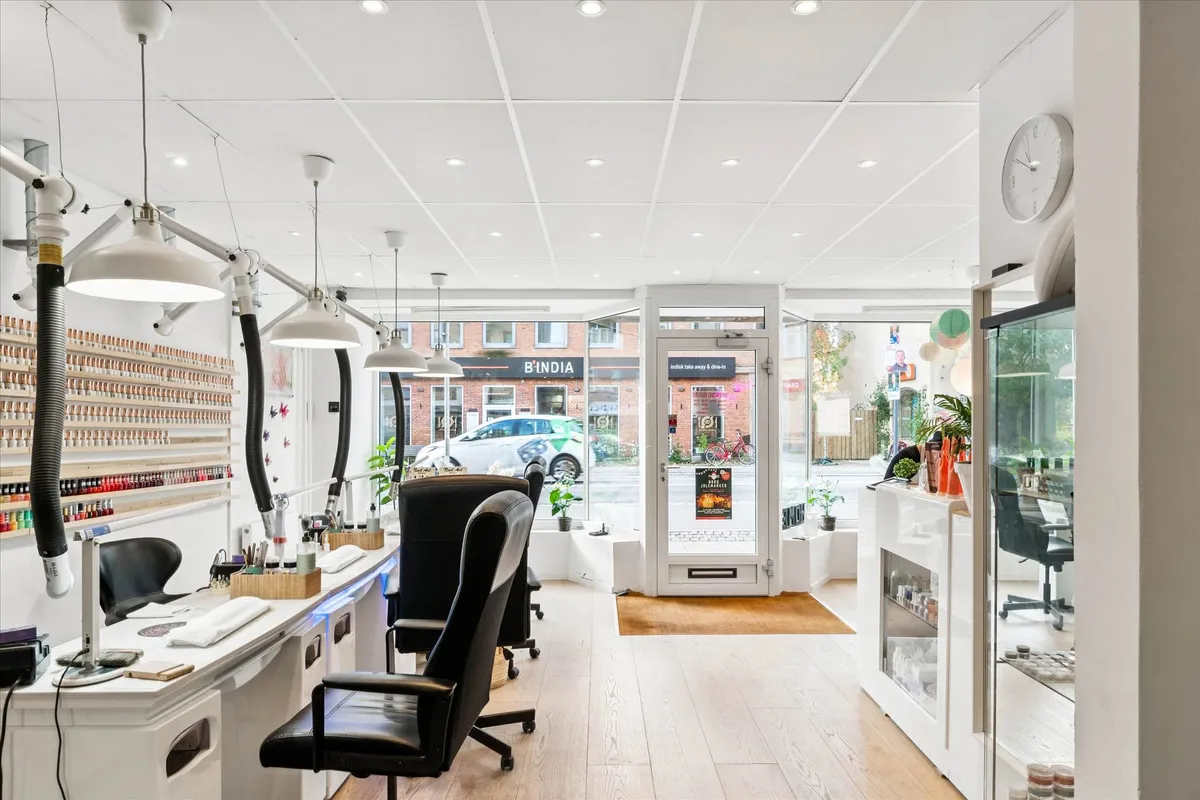 En salon med mange stole og stole med en bord og hylder med neglelak. Væggene er hvide og der er lysekroner hængende fra