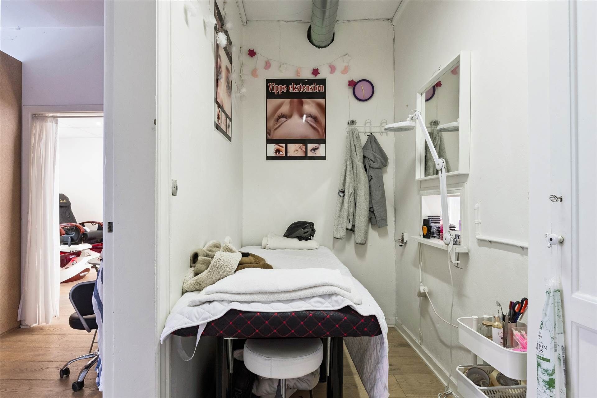 Et lille skønhedssalon med en massageseng, håndklæder, en hængelampe og en lille indgangsdør. Et sæt briller hænger på