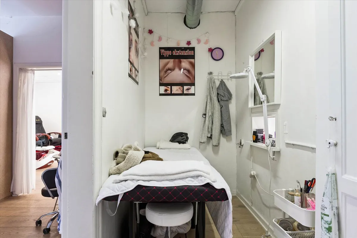 Et lille skønhedssalon med en massageseng, håndklæder, en hængelampe og en lille indgangsdør. Et sæt briller hænger på