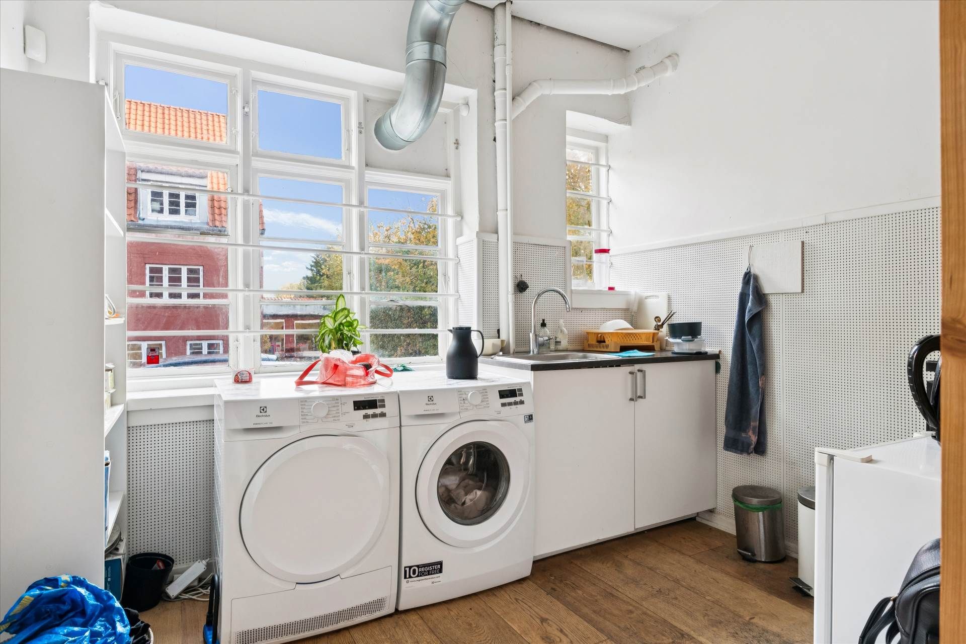 Et vaskeværelse med hvide apparater og møbler, et trægulv, og en udsigt til gaden med huse og træer