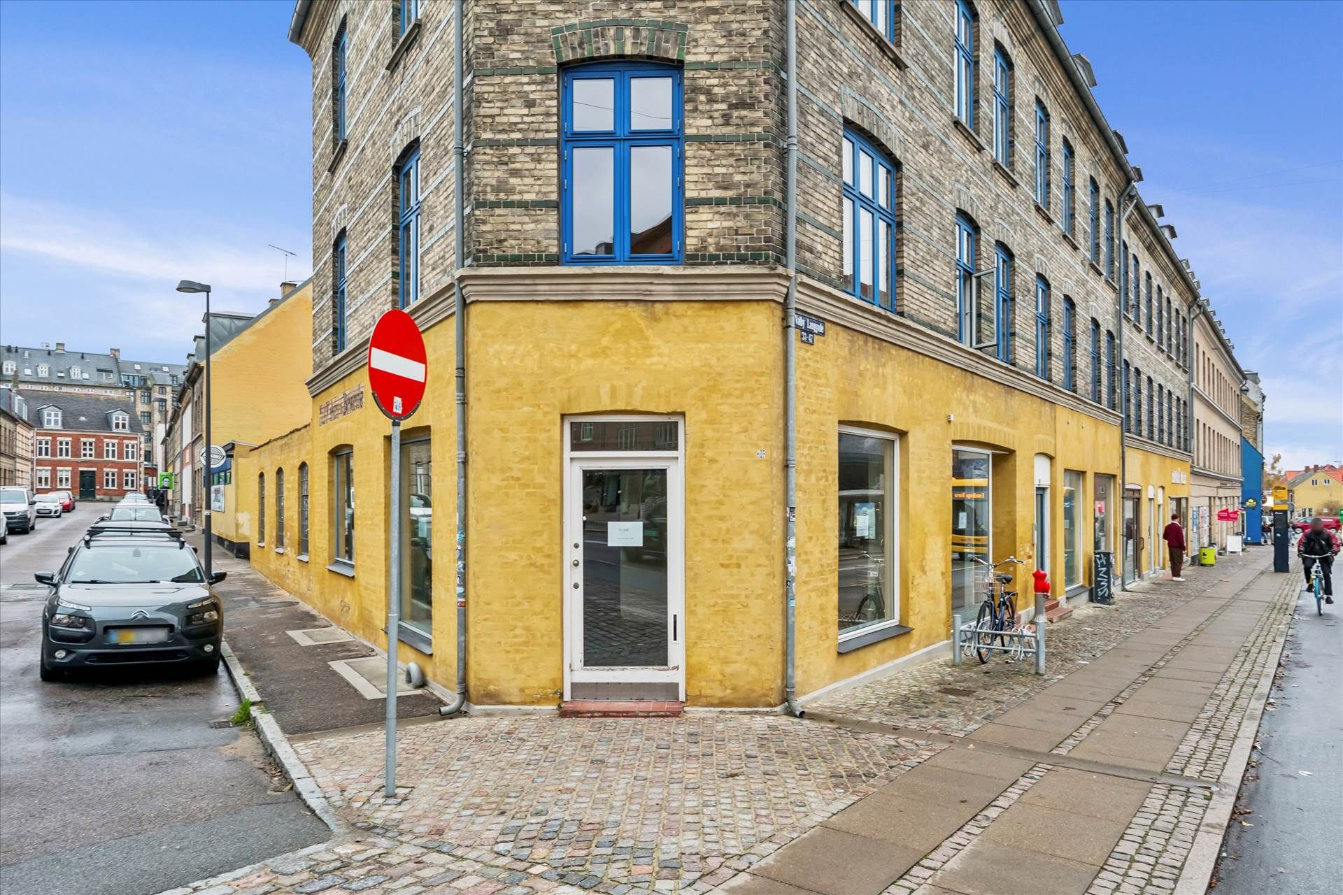 Hjørnebutik med gul facade og blå ruder på Valby Langgade med nogle cykler udenfor og en person på cykel