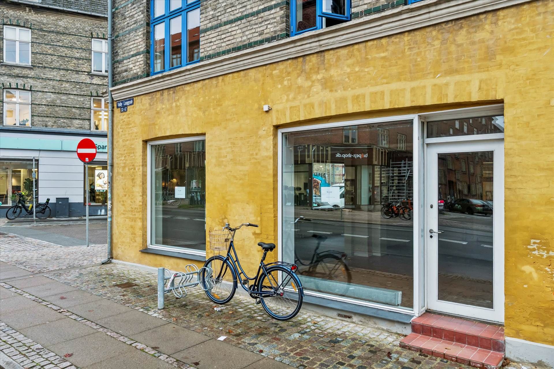 En gulemaleret hjørnebutik med glasruder og glasdør med cykelhylde og stoptegnsskilt til venstre