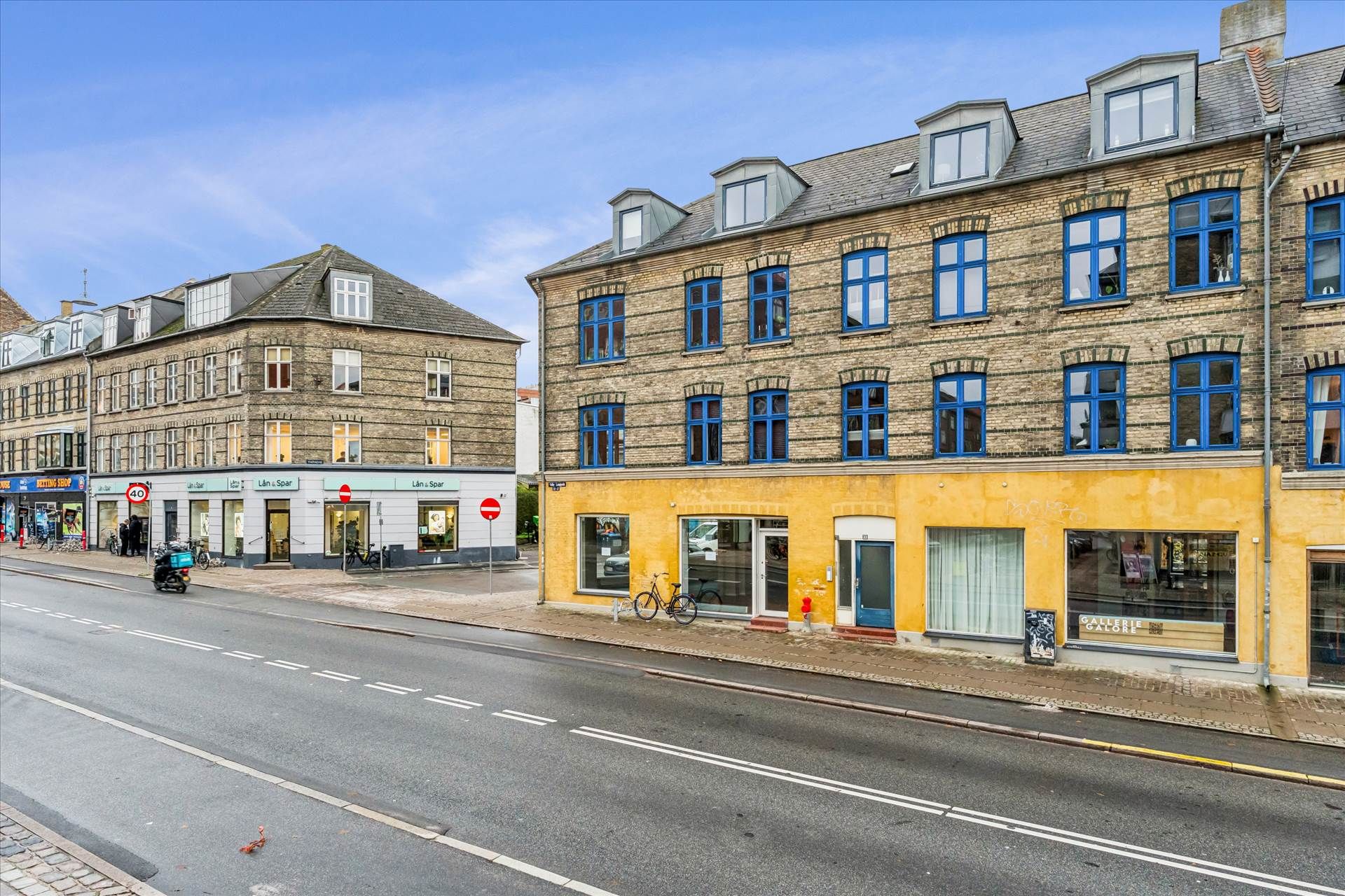 Valby Langgade med hjørnebutikker og cykler parkeret udenfor. Butikkerne er gulmalet med blå ruder og røde skilte. Der er en