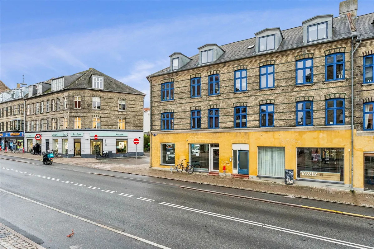 Valby Langgade med hjørnebutikker og cykler parkeret udenfor. Butikkerne er gulmalet med blå ruder og røde skilte. Der er en