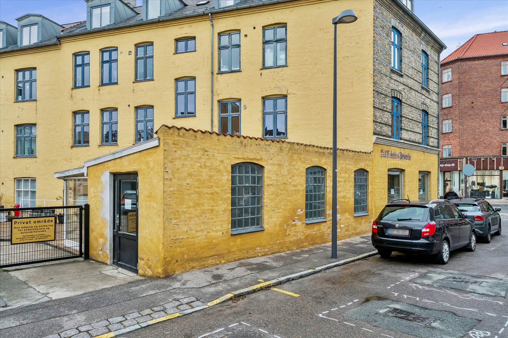 En hjørnebygning i Valby med en sort indgangsdør og en parkeringsskilt foran. To sorte biler parkeret på gaden med en søjle