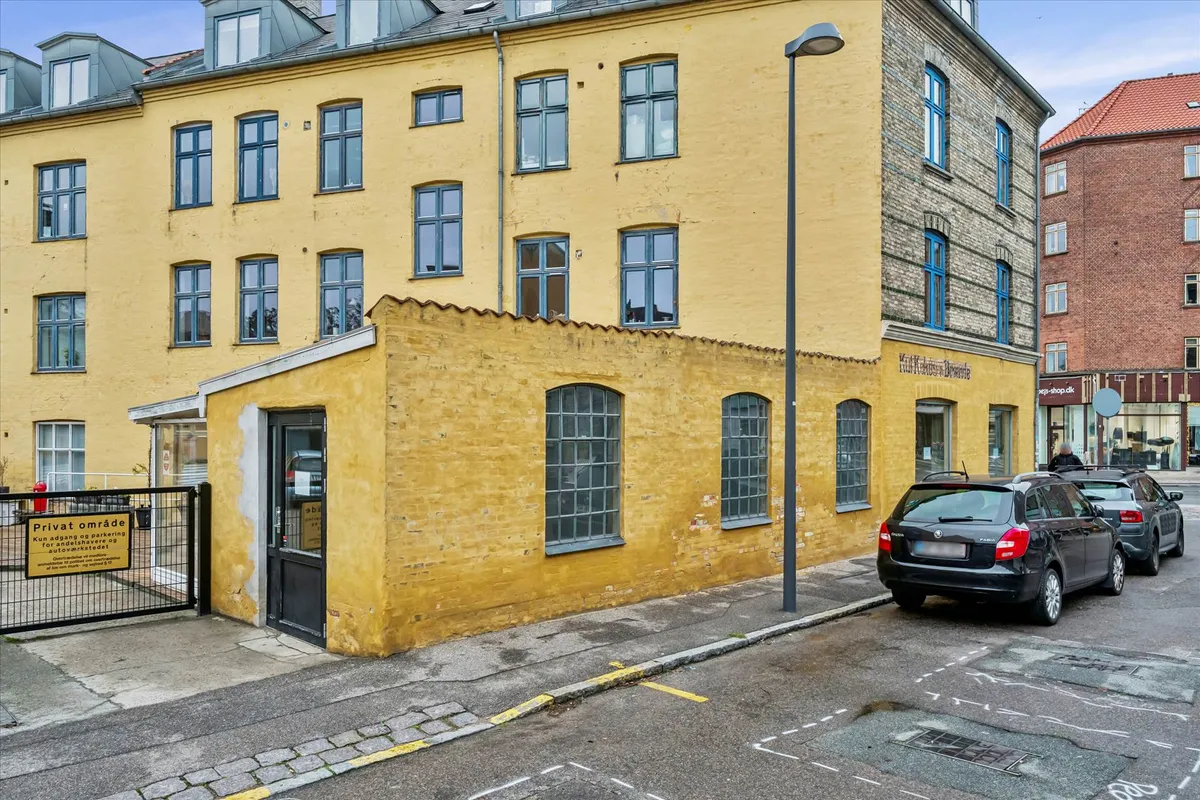 En hjørnebygning i Valby med en sort indgangsdør og en parkeringsskilt foran. To sorte biler parkeret på gaden med en søjle