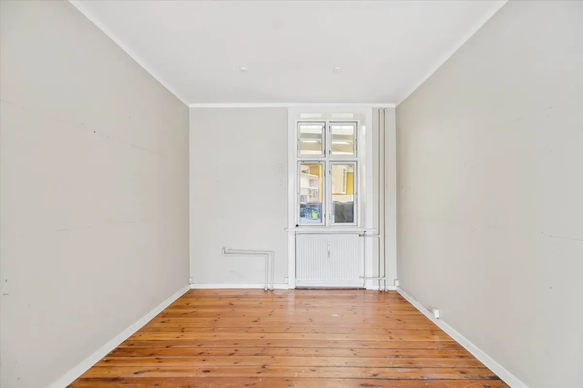 En stor, tom og lyse stue med hårdt trægulv og hvide vægge samt store vinduer mod en anden stue