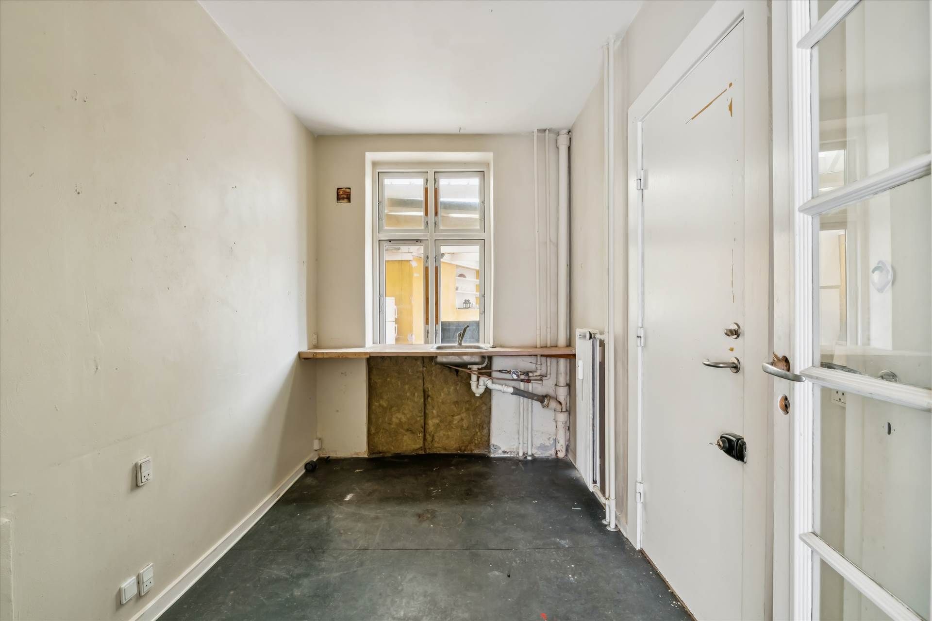 En tom hjørnebutik med tætsiddende ruder og lyse, hvide vægge. Der er et skab med en lysestage og en radiator i væggen.