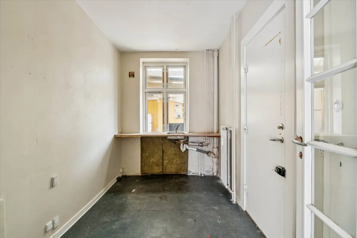 En tom hjørnebutik med tætsiddende ruder og lyse, hvide vægge. Der er et skab med en lysestage og en radiator i væggen.