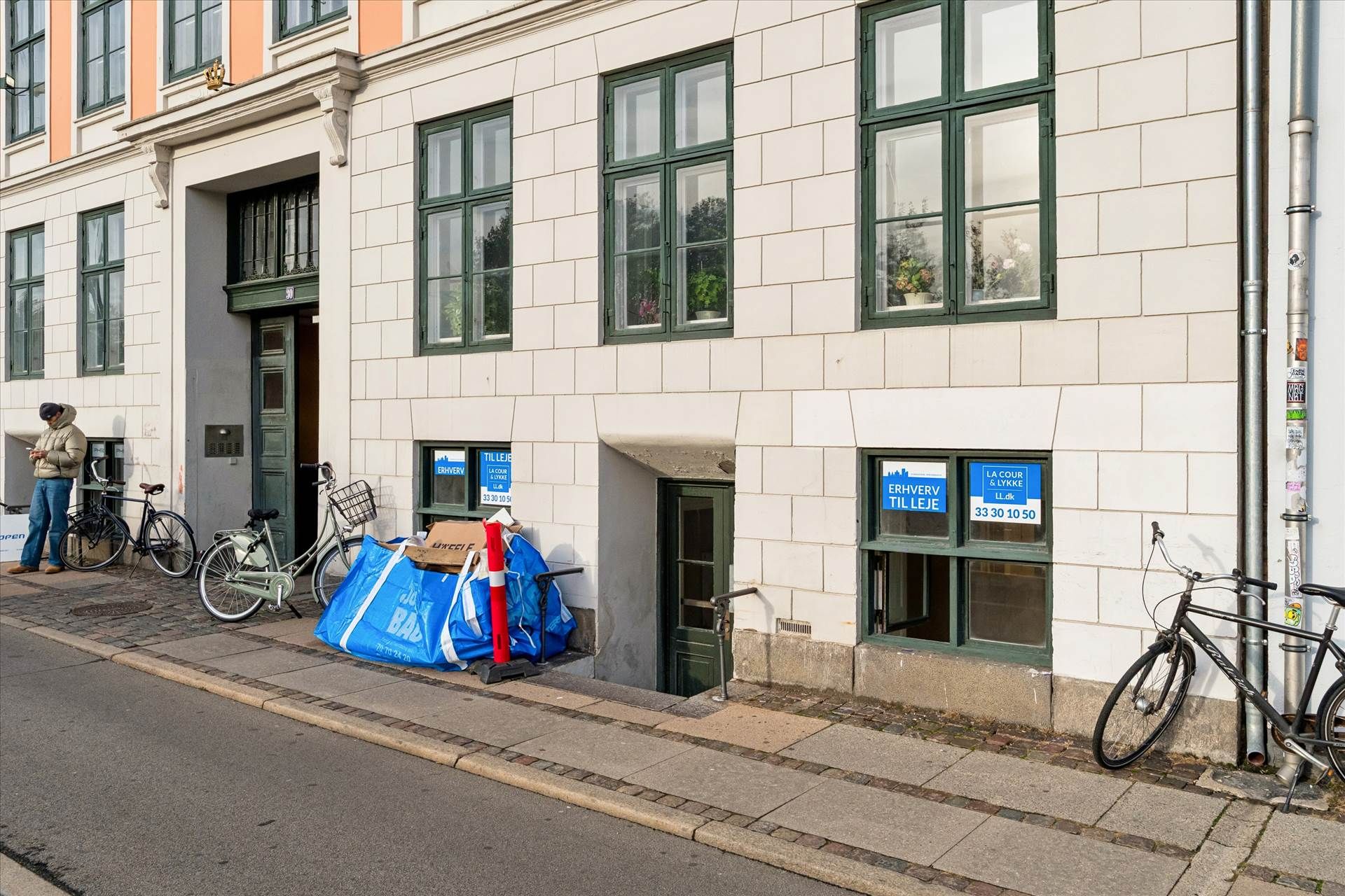 En mænd står med en jakke og læser på en avis ved en bycykelparkering på en gågade med tre cykler og et rødt og hvidt