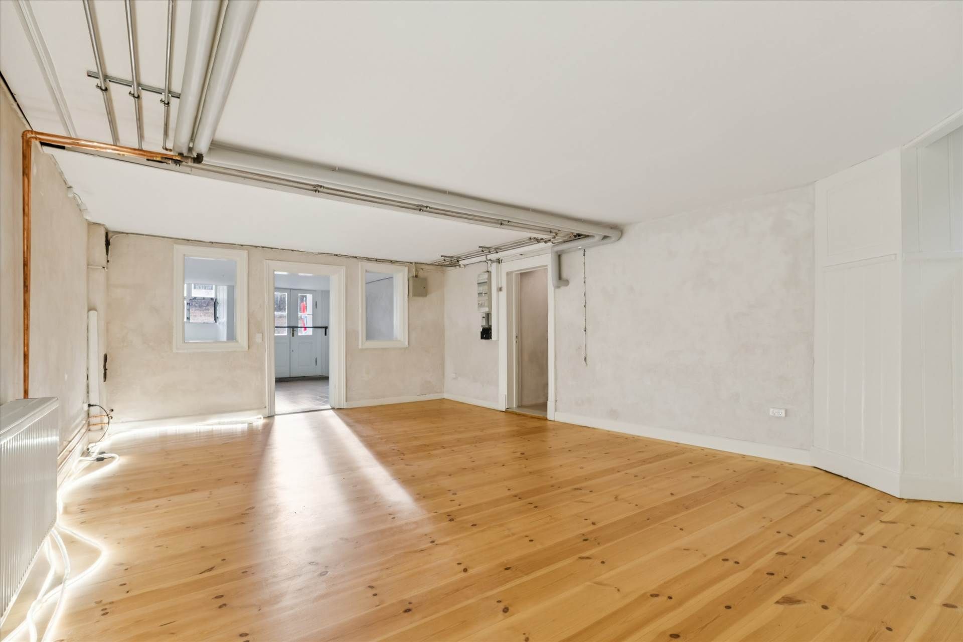 En stor tom rum med lysegult trægulv og hvide vægge samt lysekroner og radiatorer i det ene hjørne