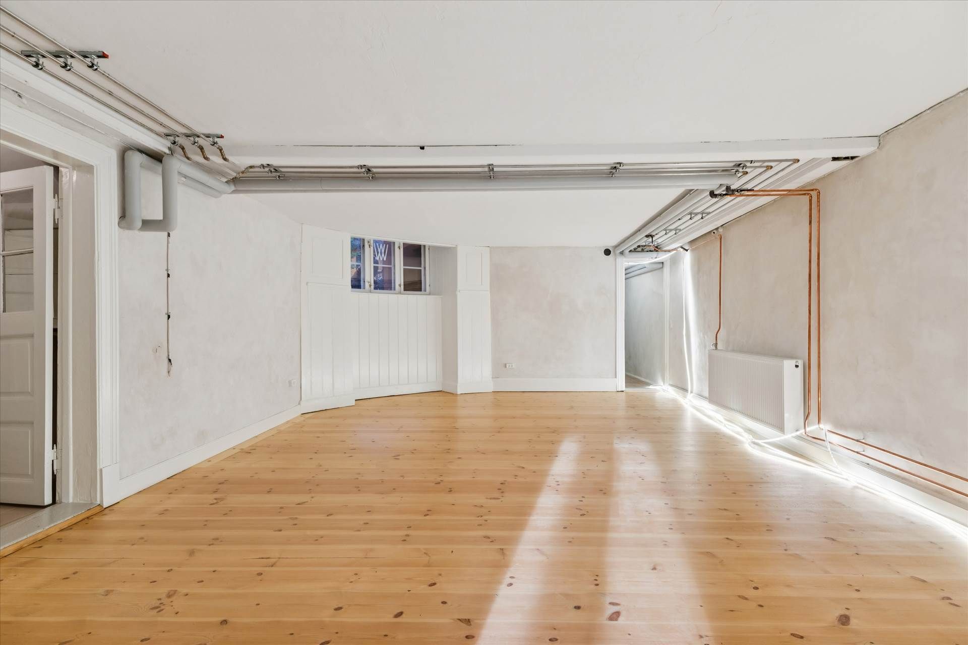 En stor, lys og velholdt indre rum med lyse trægulve og hvide vægge, udstyret med moderne radiatorer og ventiler