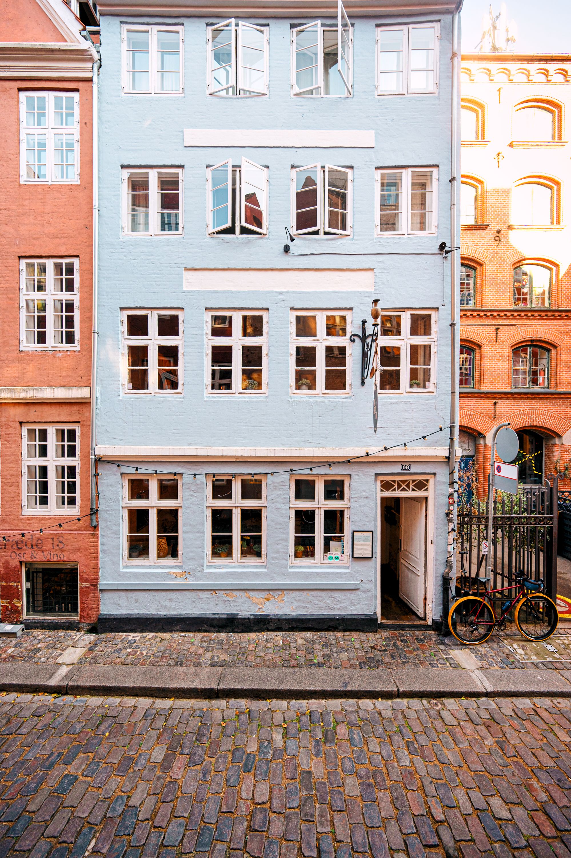 En charmerende restaurant med åbne vinduer og lyse lysekroner. Københavnerhus med hvide vindueskarme og røde mursten. En cykel står parkeret foran restaurantdøren.