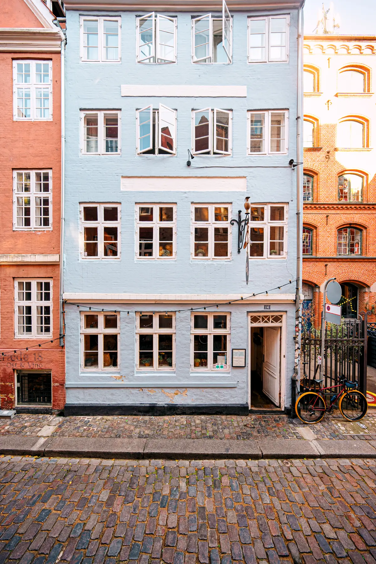 En charmerende restaurant med åbne vinduer og lyse lysekroner. Københavnerhus med hvide vindueskarme og røde mursten. En cykel står parkeret foran restaurantdøren.