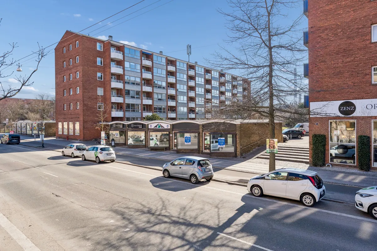 En stor bygning med flere lejligheder ligger ved siden af en gågade med parkerede biler og grønne planter og træer
