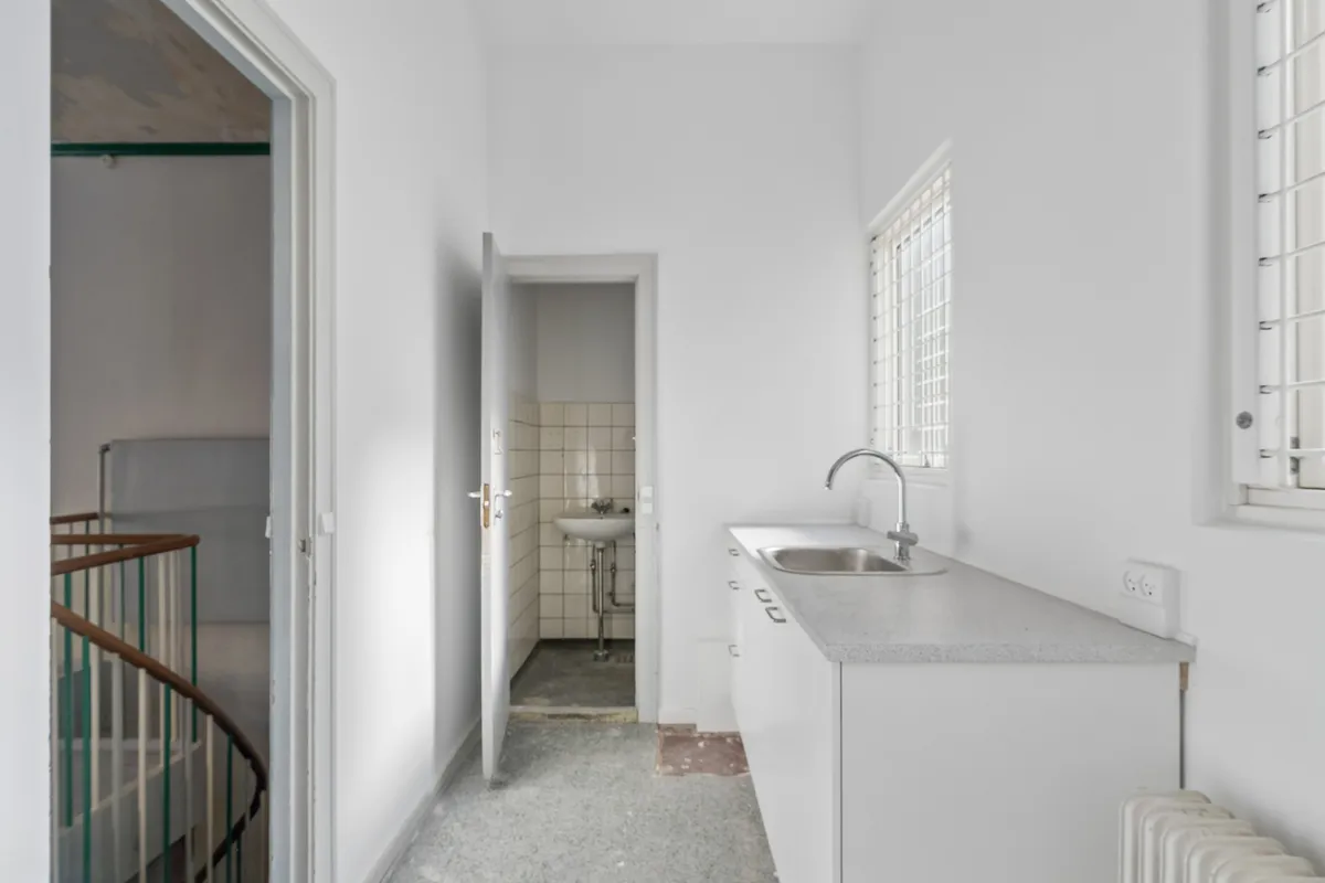 Kontor til leje med lyse, rene værelser og hvidt køkken med rustfrit stålvask og vandhaner samt en træspiraltrappe i baggrunden