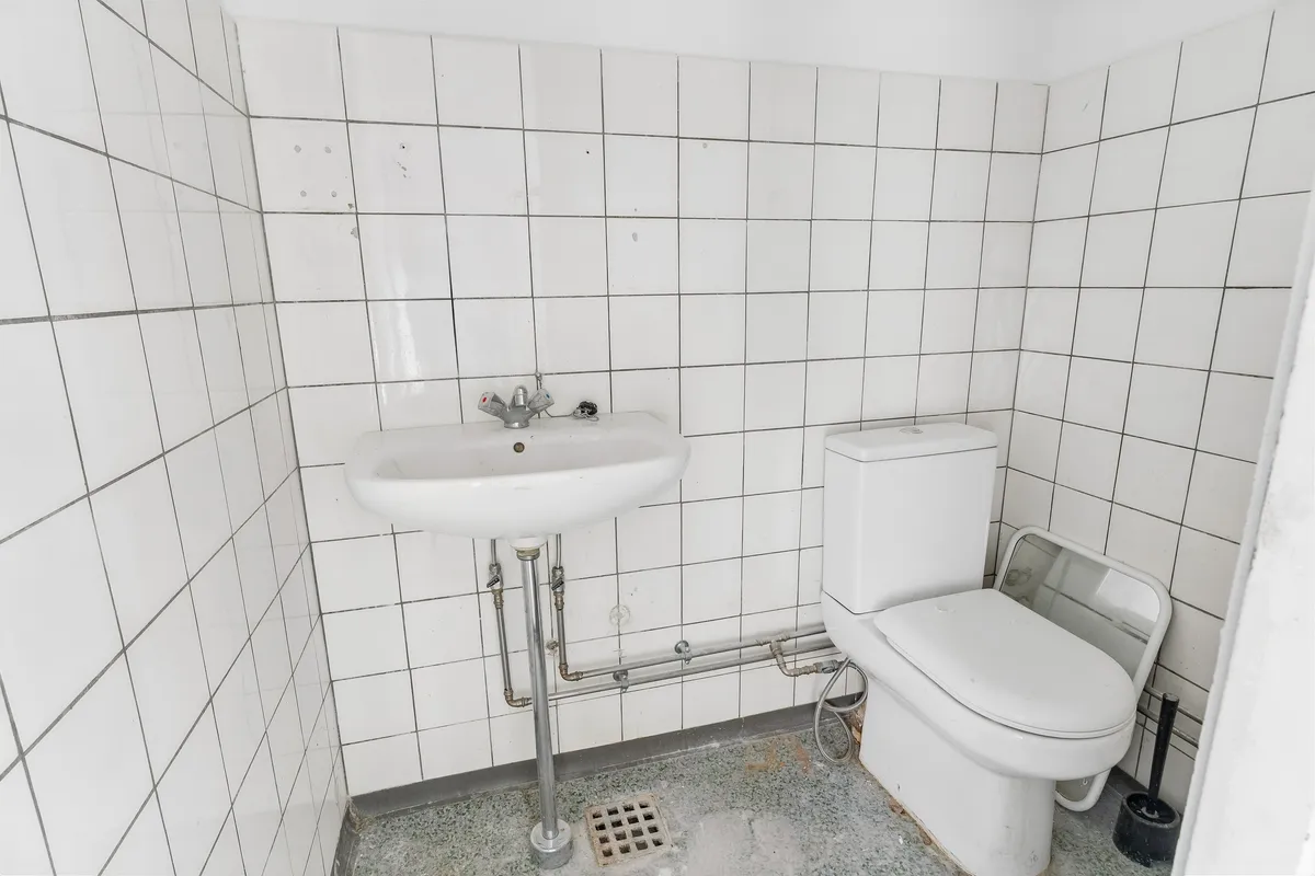 Hjørnetoilet med hvid toiletvogn, hvid keramik og metalforstærkning. Tæt ved en hvid vask med kromfarvet vandhane og rørforbindelser. Rundt om er der hvide fliser med grå og lyseblå kant.