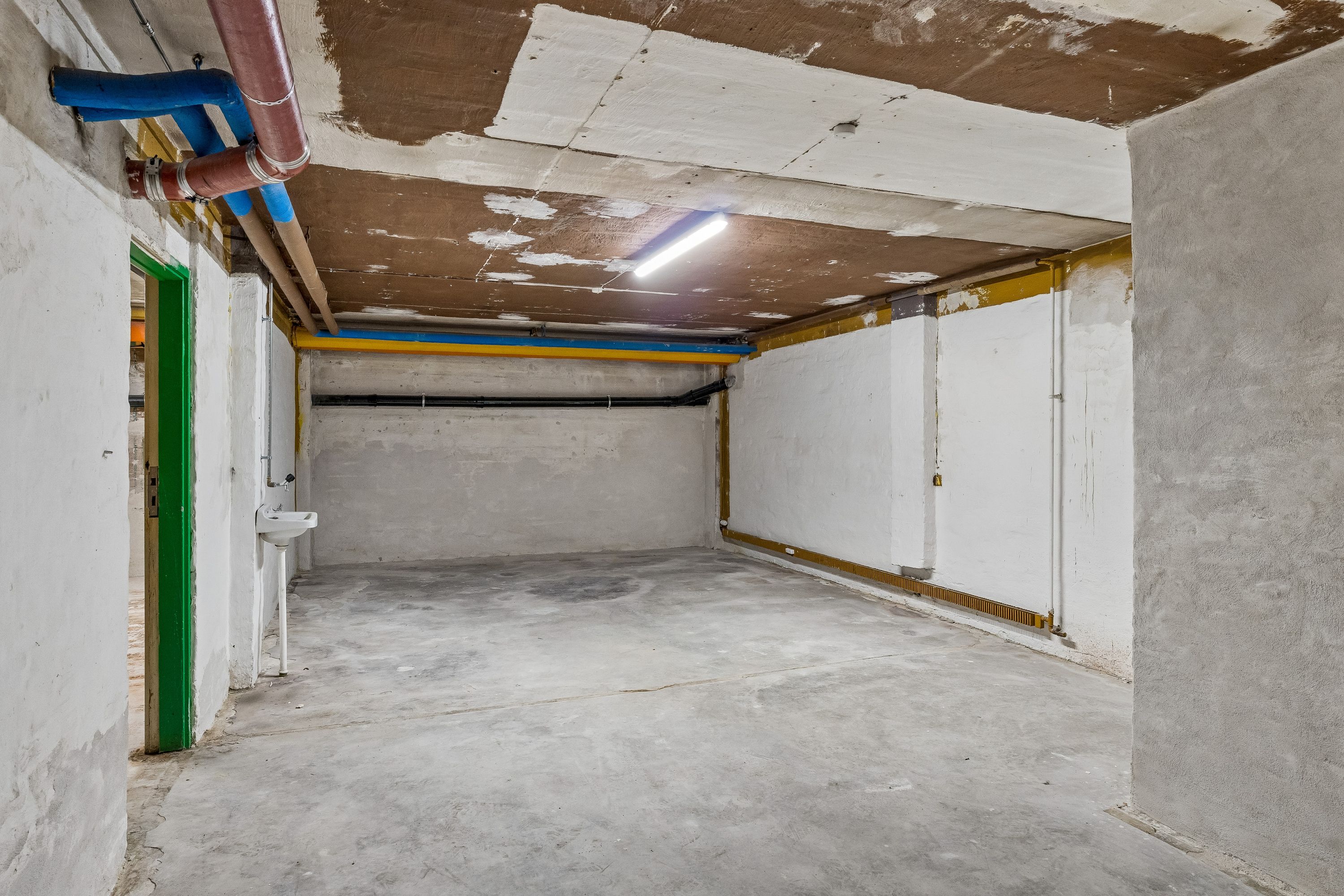 En stor og lys kælder med cementgulv og lyse, lysegrå vægge, der er til leje. Der er en åben dør og en væskehåndvask med vandhane. Loftet har hængende rør og en lysskiltet.