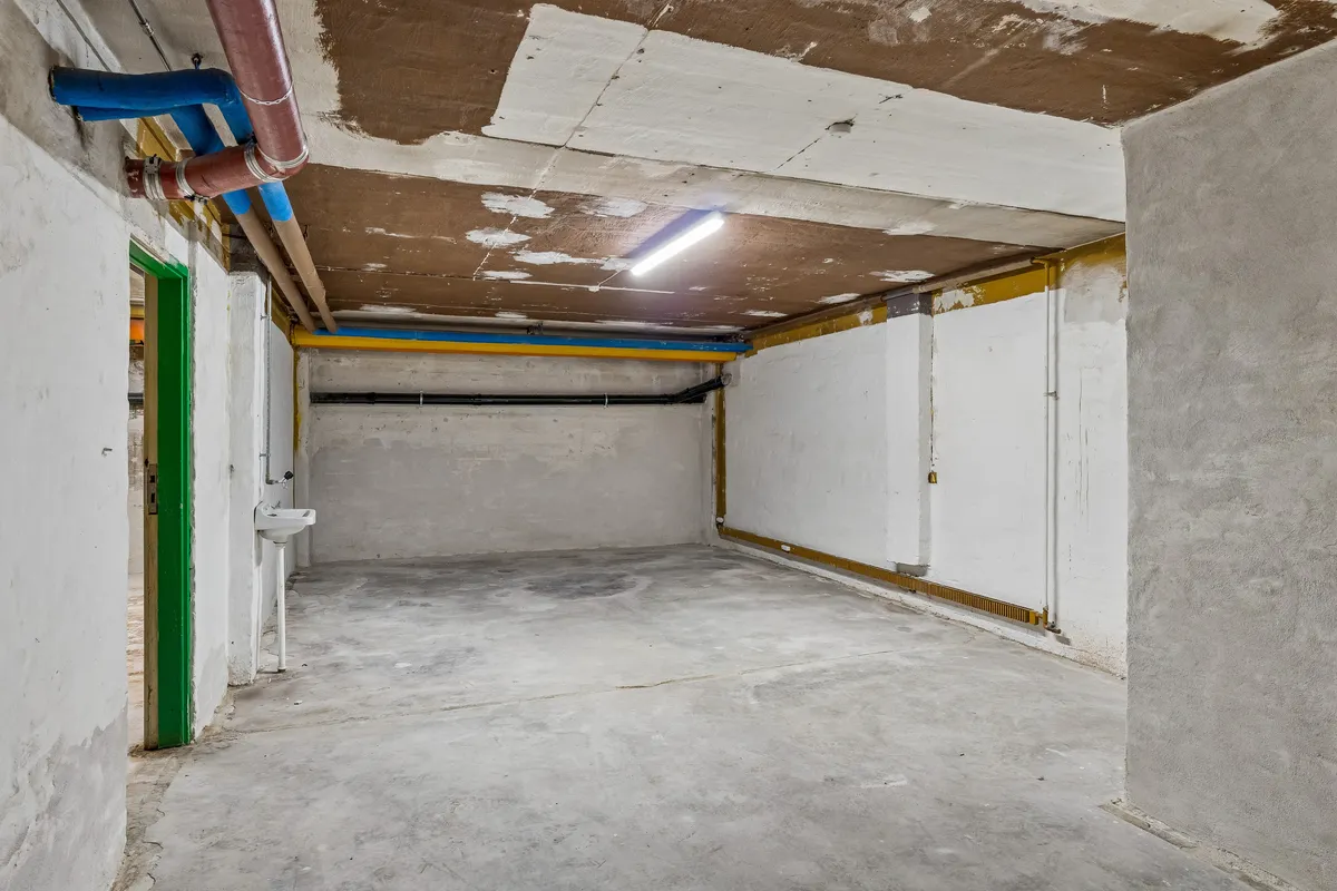 En stor og lys kælder med cementgulv og lyse, lysegrå vægge, der er til leje. Der er en åben dør og en væskehåndvask med vandhane. Loftet har hængende rør og en lysskiltet.