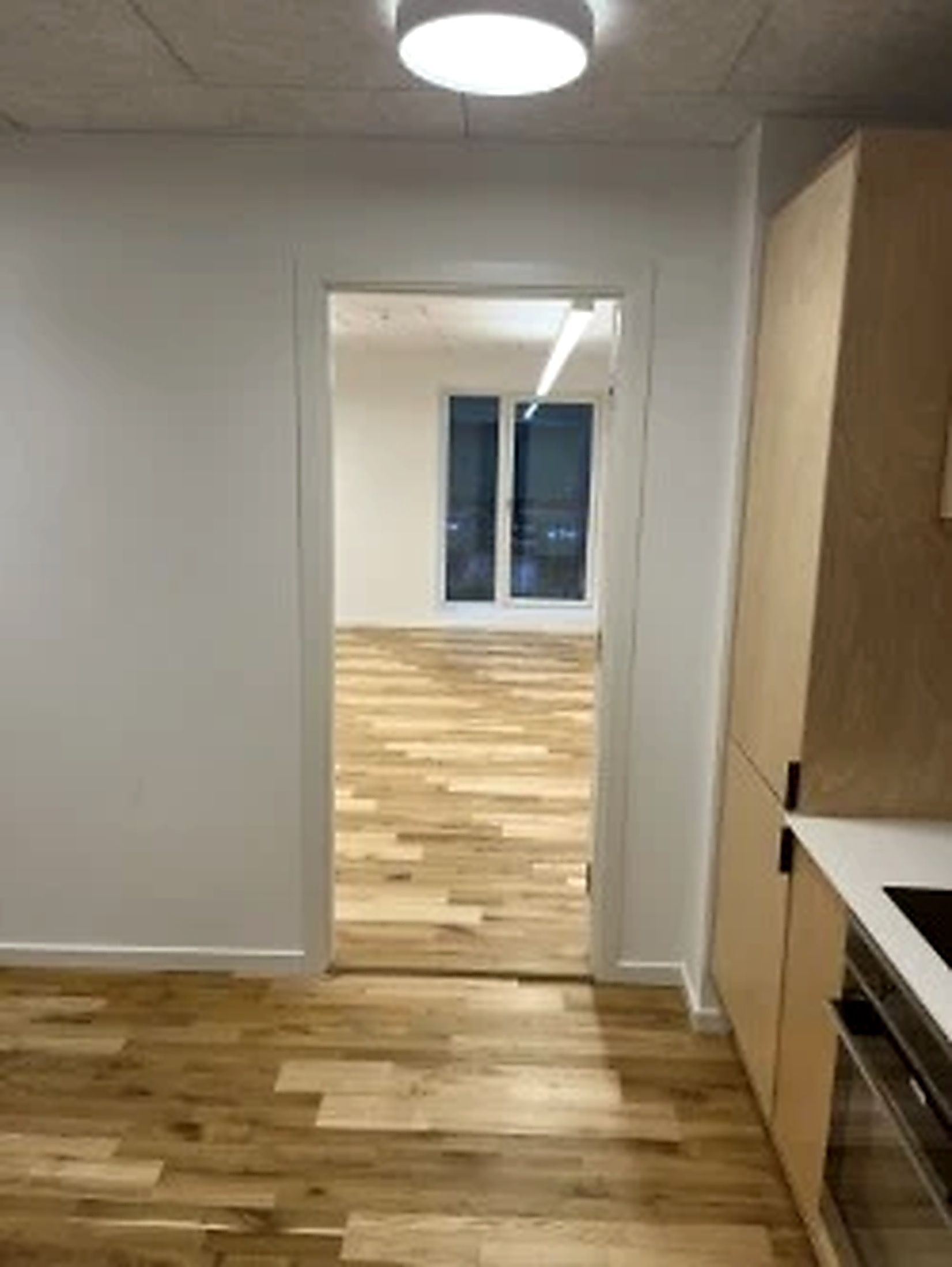 Kontorrum med åbne døre og lyse loftslamper. Gulvbelægning af træ og lyse vægge. Møbler i lysebrunt og hvidt. Et køkken med en indbygget ovn og en spisestue.