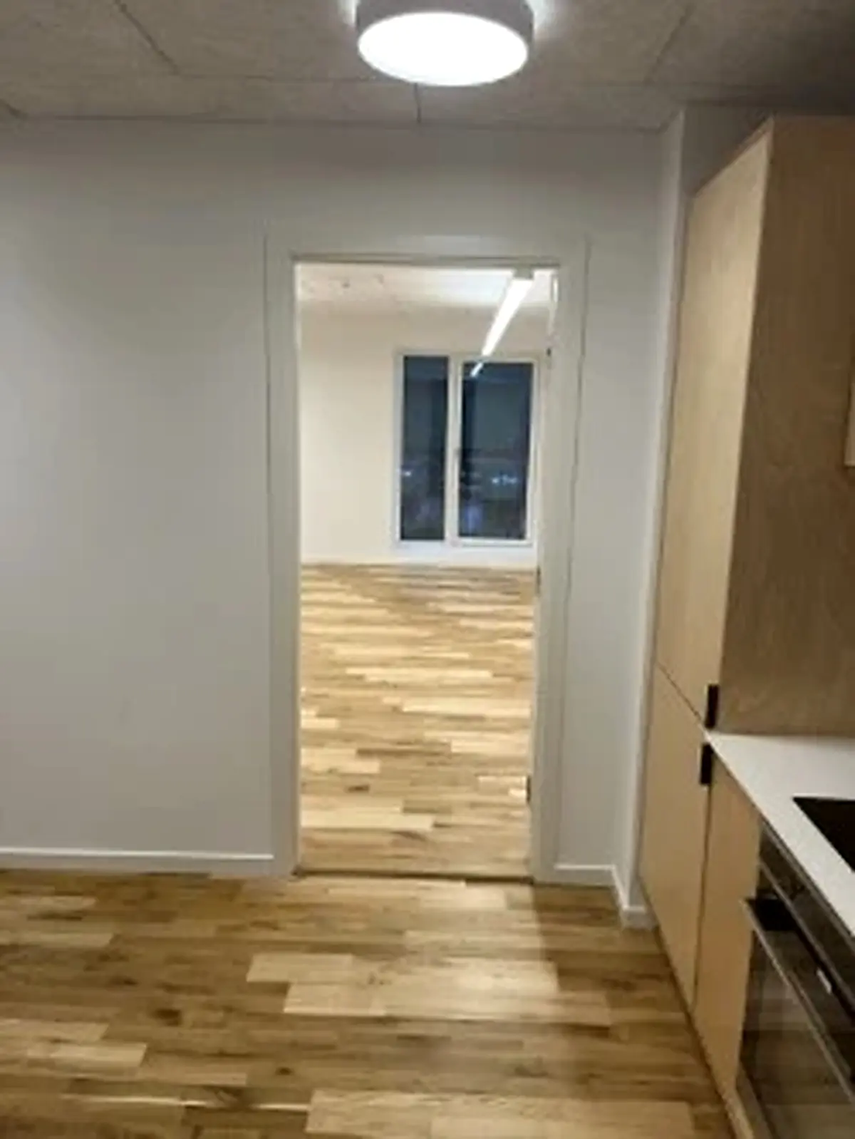 Kontorrum med åbne døre og lyse loftslamper. Gulvbelægning af træ og lyse vægge. Møbler i lysebrunt og hvidt. Et køkken med en indbygget ovn og en spisestue.