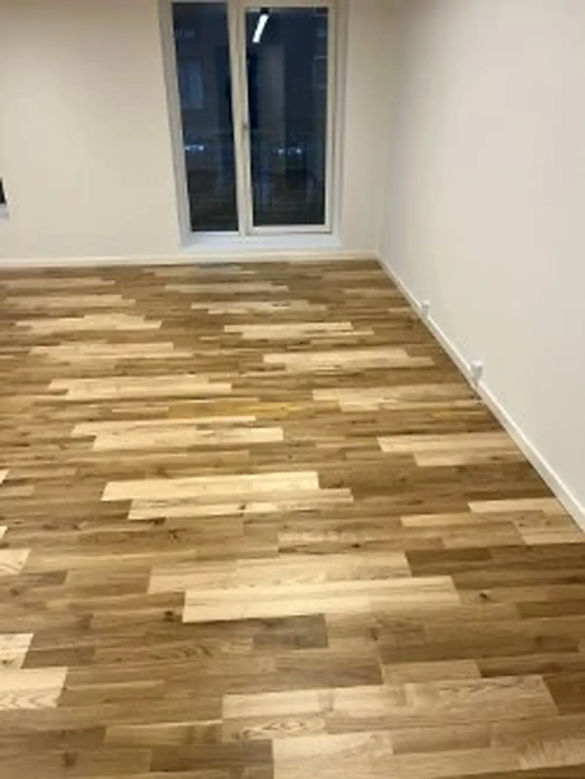 Et tomt kontorrum med hårdt trægulv og glasdøre til et andet værelse. Gulvet er i hvidt og brunt, og der er en hvid væg med et stikkontakt til højre.