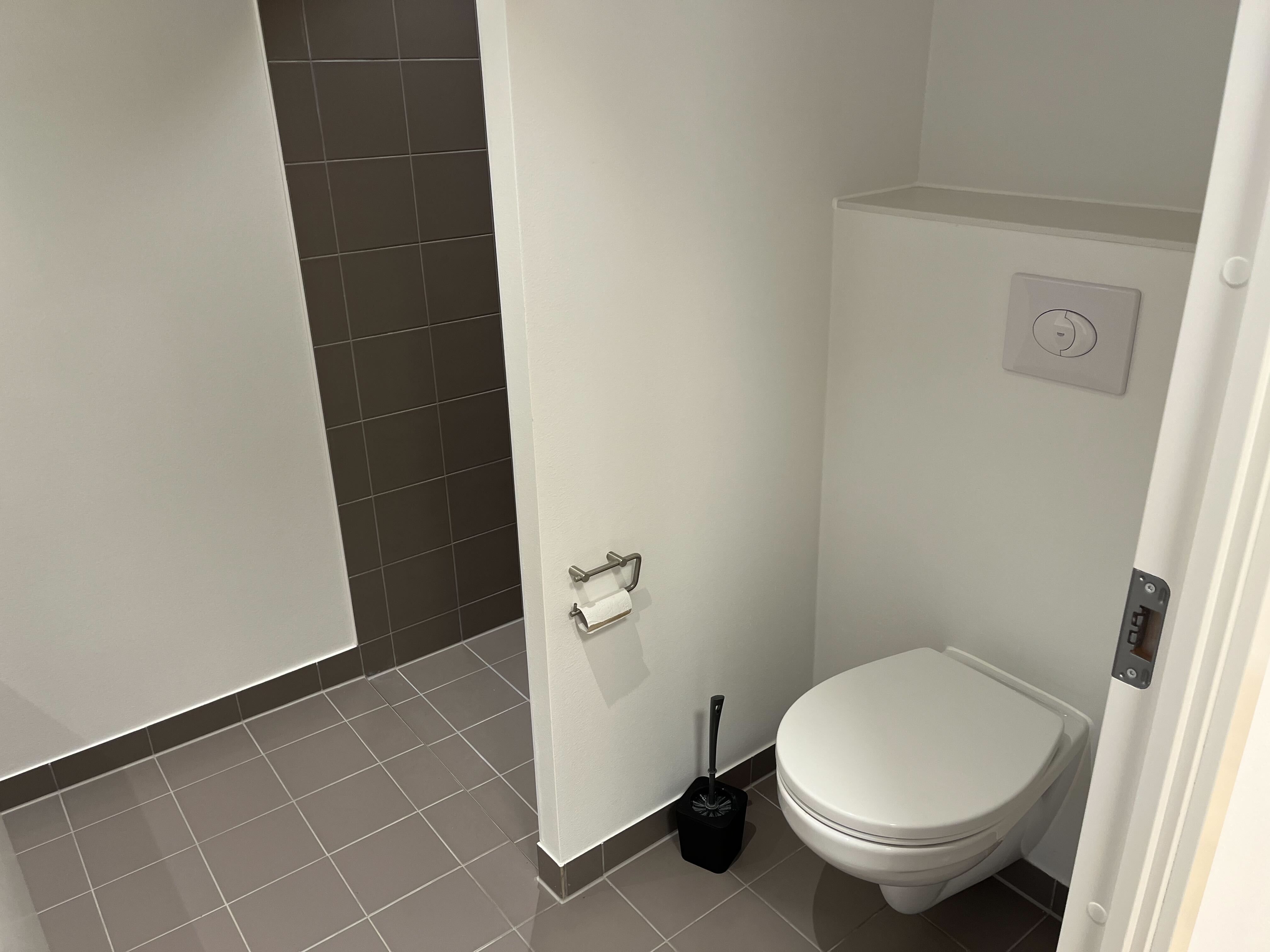 Lyst og funktionelt kontorlokale med toilet, bad og køkken. Toilet med hvid toiletbruse og hængende toiletpapir. Moderne og rent gulv med grå flise.