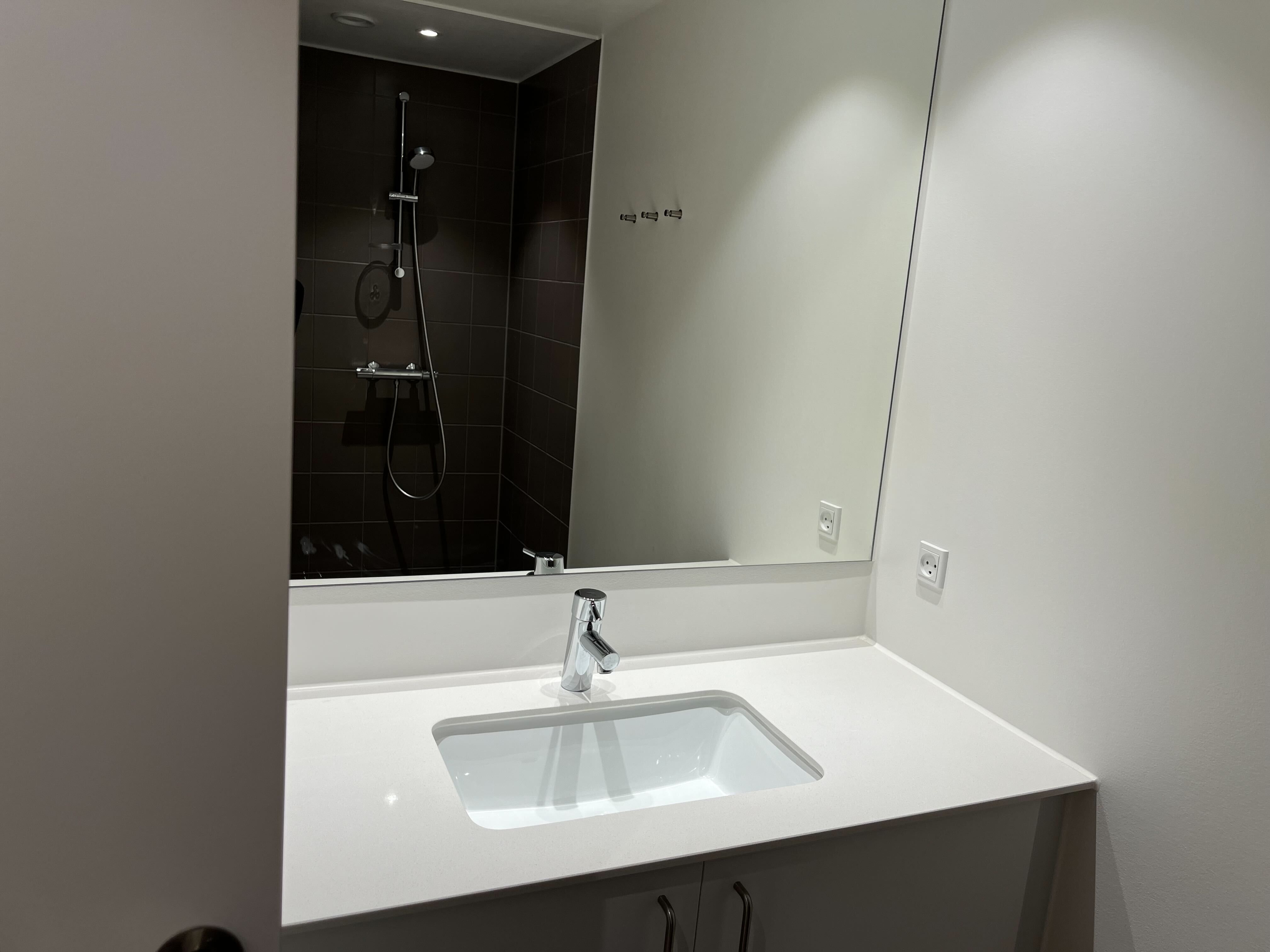 Rent kontor med hvidt toilet og sort badeværelse. Toiletspejl med refleksion af brusebad og hvid vask. På væggen er der et hvidt og et gråt stikkontakt.