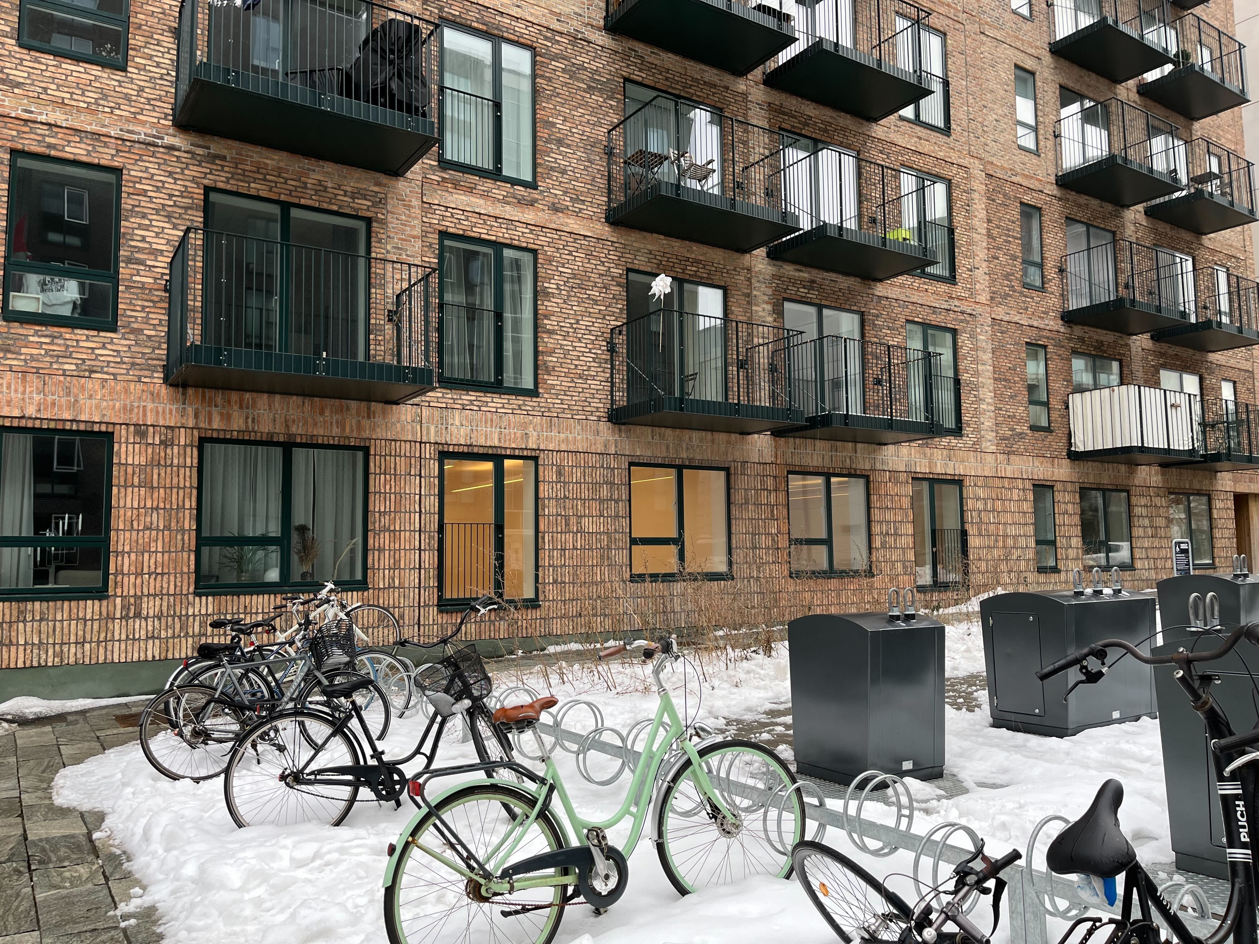 En række højhuslejligheder med balkoner og udsigt til grønne områder. Få cykler parkeret foran et hus med høje, højrekantede vinduer og mange balkoner