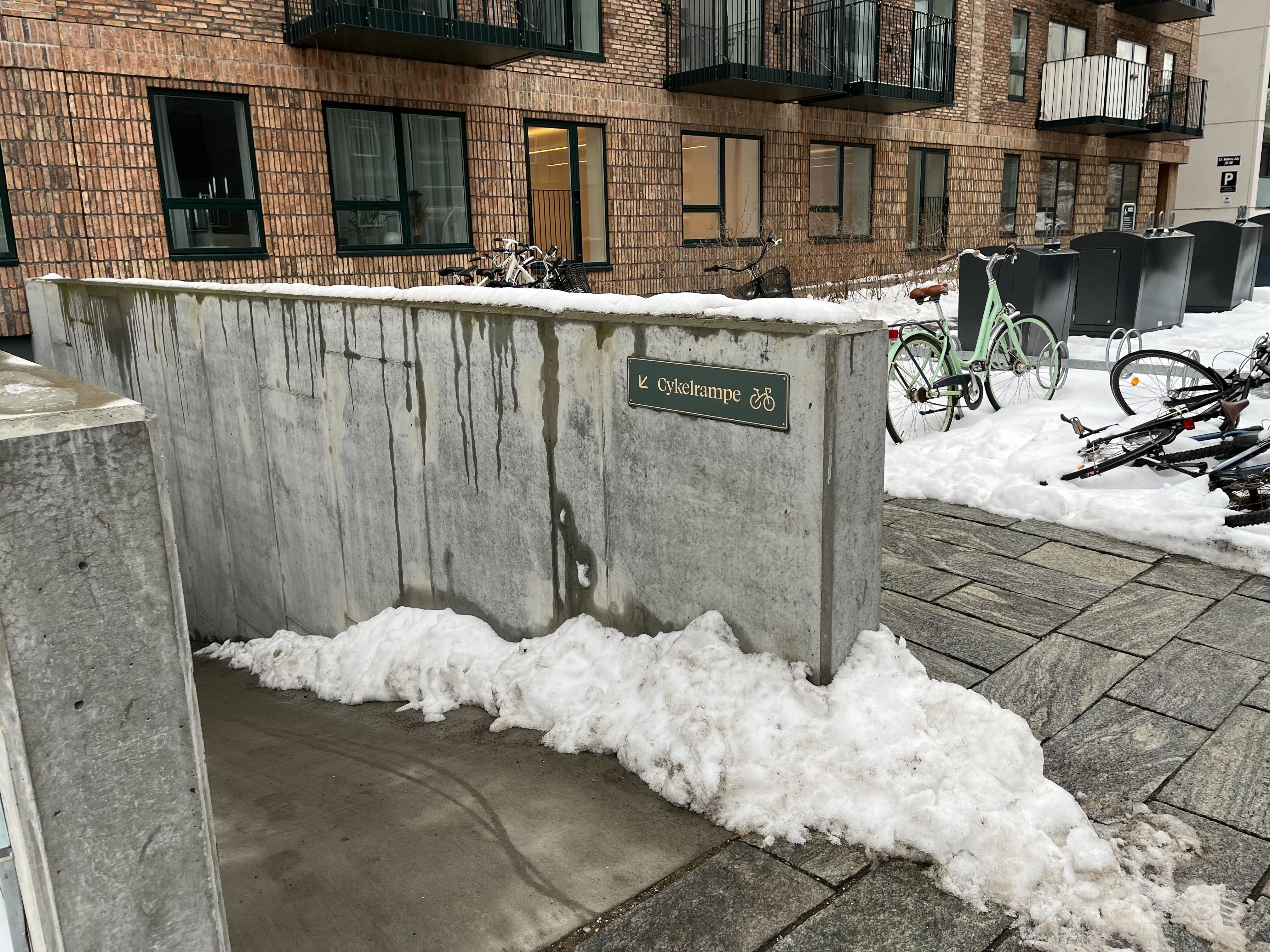 Mur med skilt Cykelråmpe og vinterligt landskab med snepletter og parkerede cykler foran et lejlighedskompleks med balkoner og røde mursten