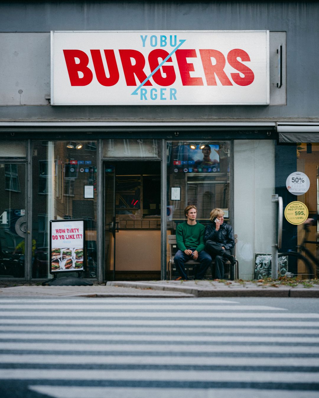 Par sidder på en stol foran burgerrestauranten Yobu Burgers på Østerbrogade. Der er skilte med burgerbilleder og teksten "How dirty do yo like it?" og "50%".