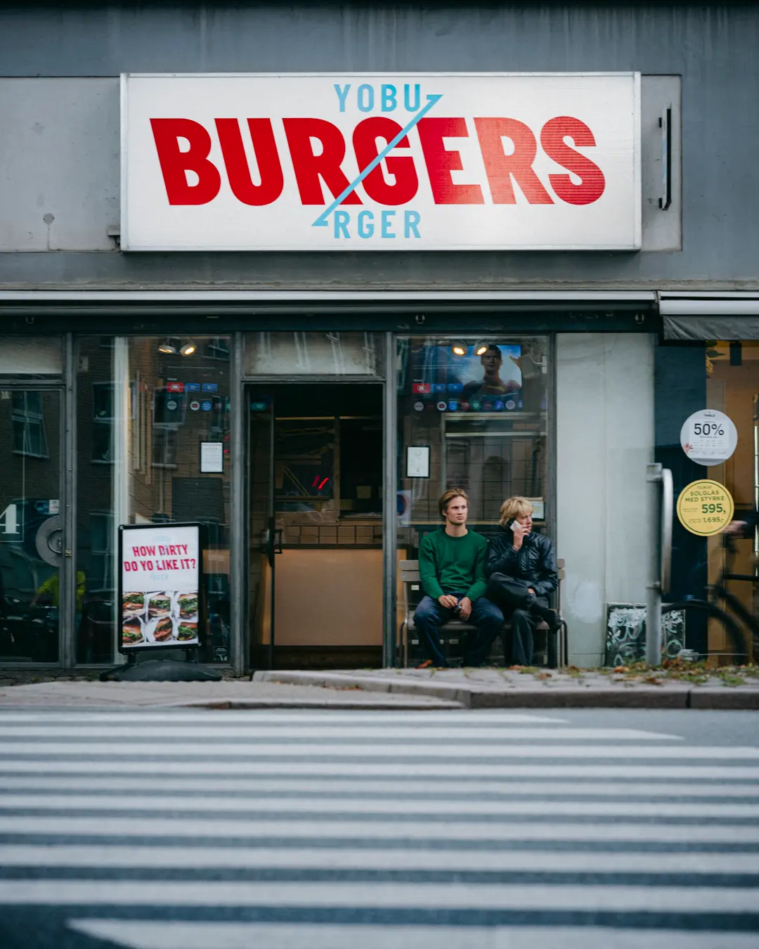 Par sidder på en stol foran burgerrestauranten Yobu Burgers på Østerbrogade. Der er skilte med burgerbilleder og teksten "How dirty do yo like it?" og "50%".