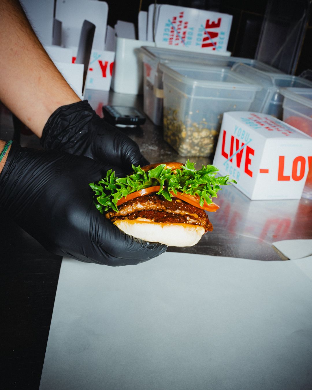 Person iført sort handske holder take-away burger med krydret kød og frisk salat og tomat i hånden