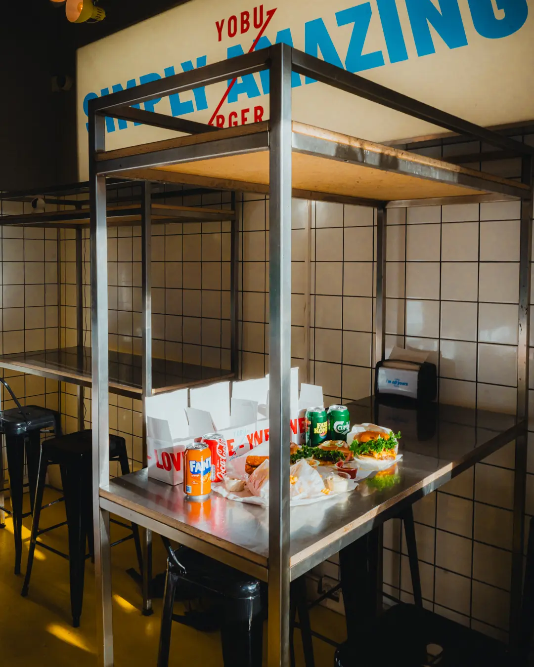 Take-away med burger, fanta og cola på metalbord i lokal med krydsfiner og kakeloverflade. Lokal med opskriften "Simply Amazing Burger" på væggen.
