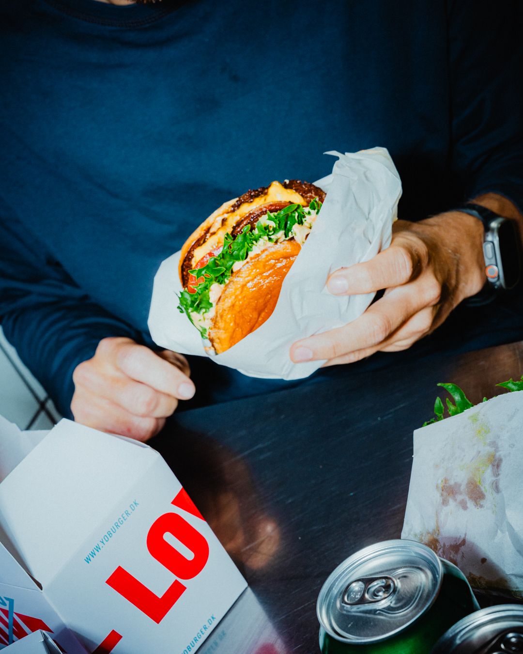 En mand holder en burger med grøn salat og andre ingredienser i hånden og sidder ved et bord med to dåser