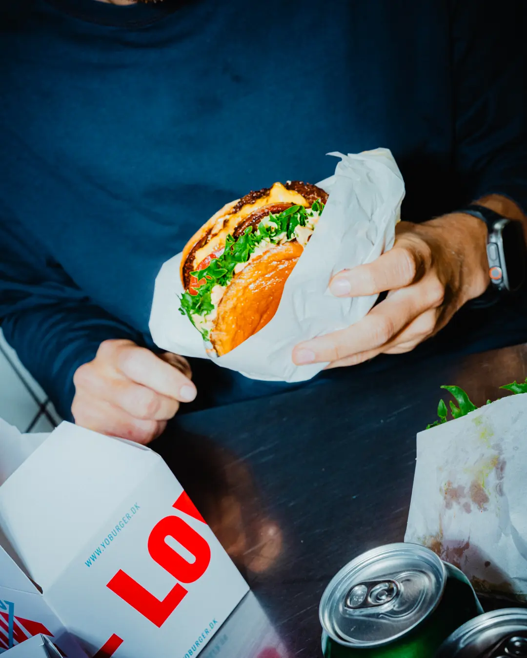 En mand holder en burger med grøn salat og andre ingredienser i hånden og sidder ved et bord med to dåser