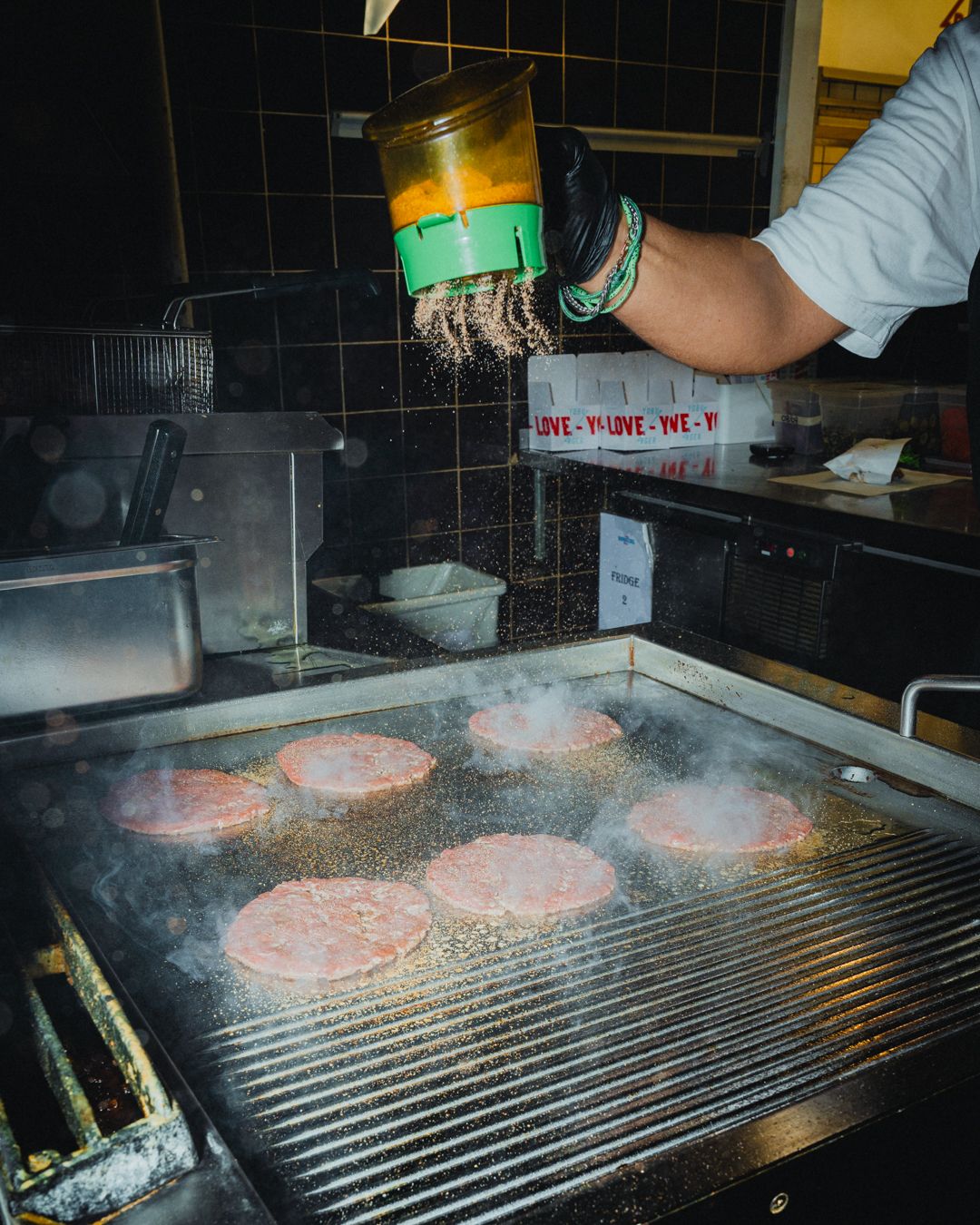 En person i en hvid skjorte og sorte handsker sprøjter krydderier over hamburgere, der koges på en grill i et moderne køkken med lysegul baggrund