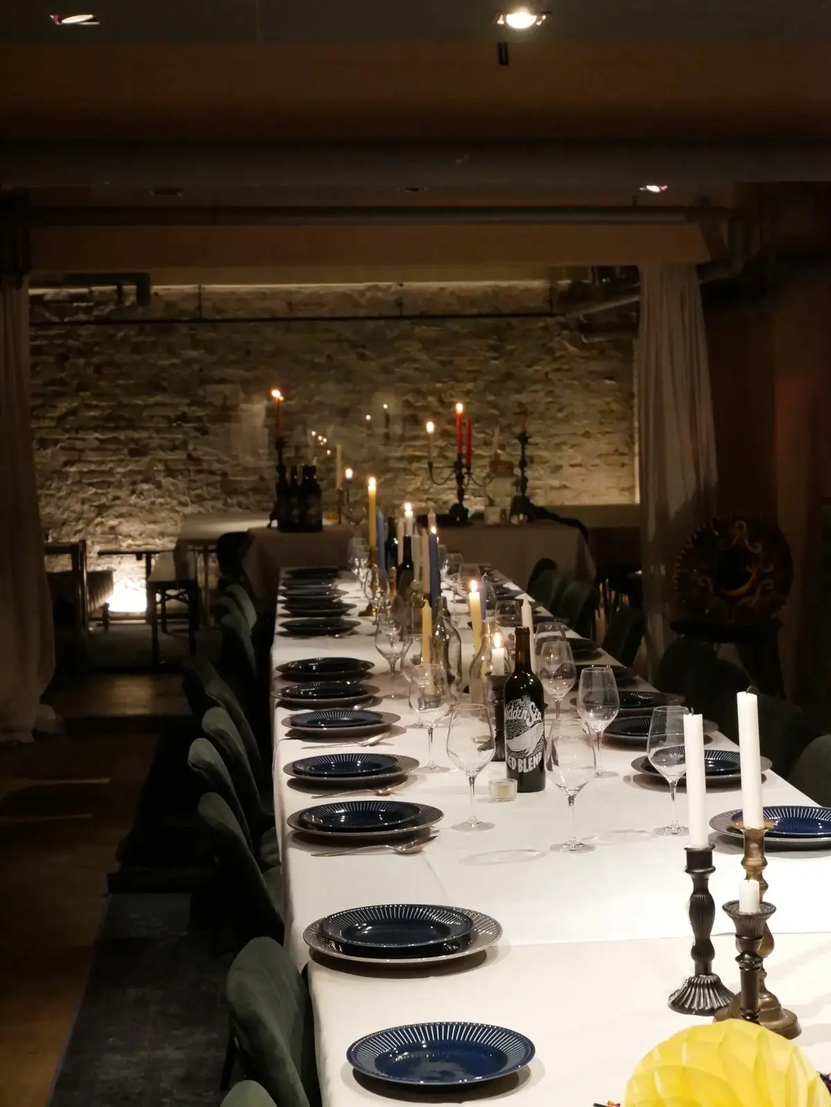 En lang bord med bordvin, hvidt linne og lysestager med brændende stearinlys og vinflasker placeret i en restaurant med stenmur og mørkt interiør