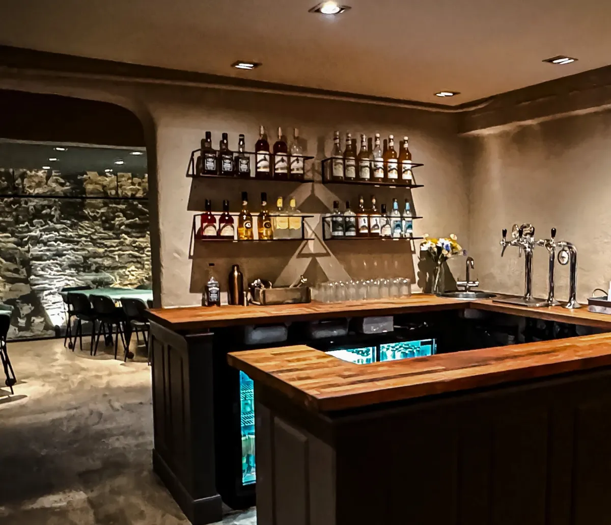 Take-away-bar med udsigt til stenmur og højt loft, to bardiske med udvalg af drikkevarer og glas samt serveringsudstyr