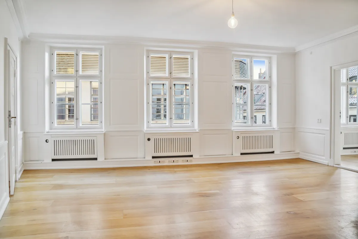 Kontor med højloft og lyse trægulve. Vinduer med hvide luder, hvide vægge og hvide radiatorer. Lampe med lyspære og to døre i hvidt.