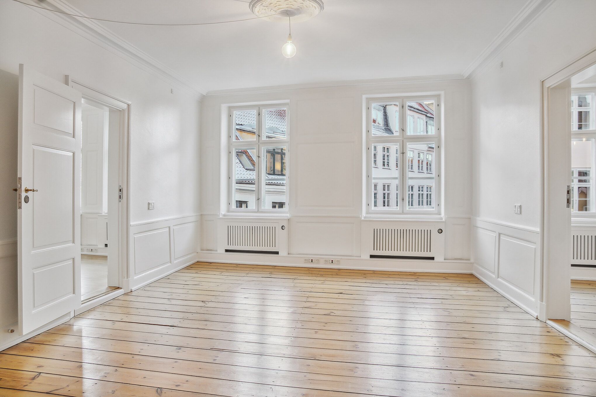 Stort, lyseværelse med højloft og trægulv, to vinduer, hvide vægge, lysekroner, radiatorer og åbne døre til andre værelser