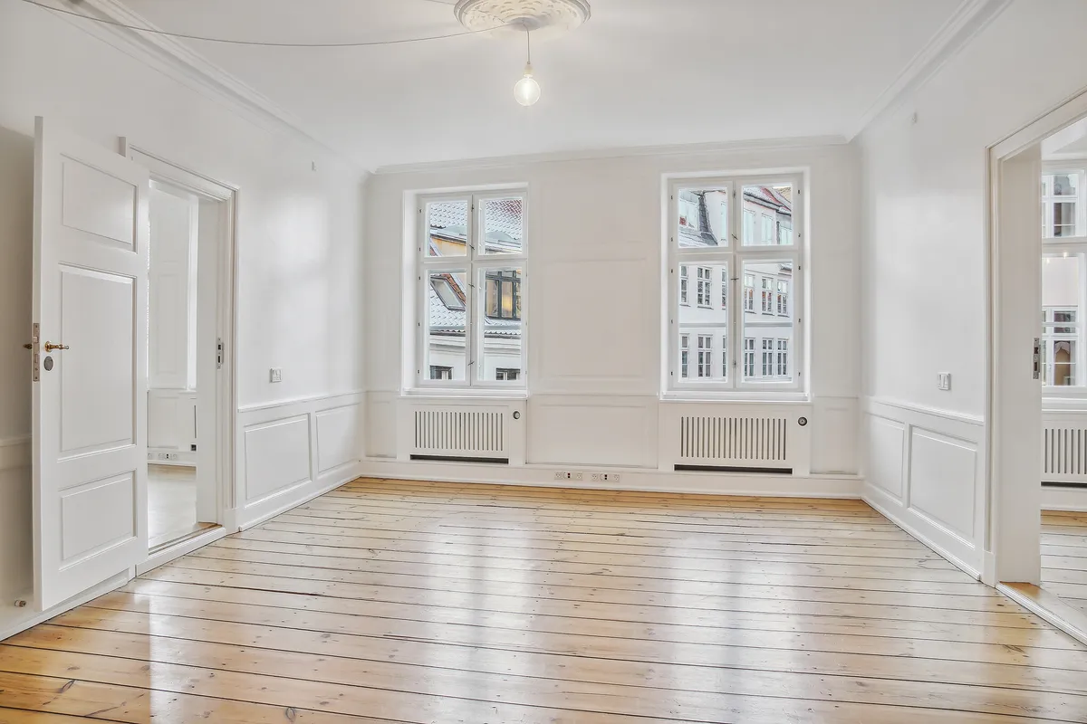 Stort, lyseværelse med højloft og trægulv, to vinduer, hvide vægge, lysekroner, radiatorer og åbne døre til andre værelser