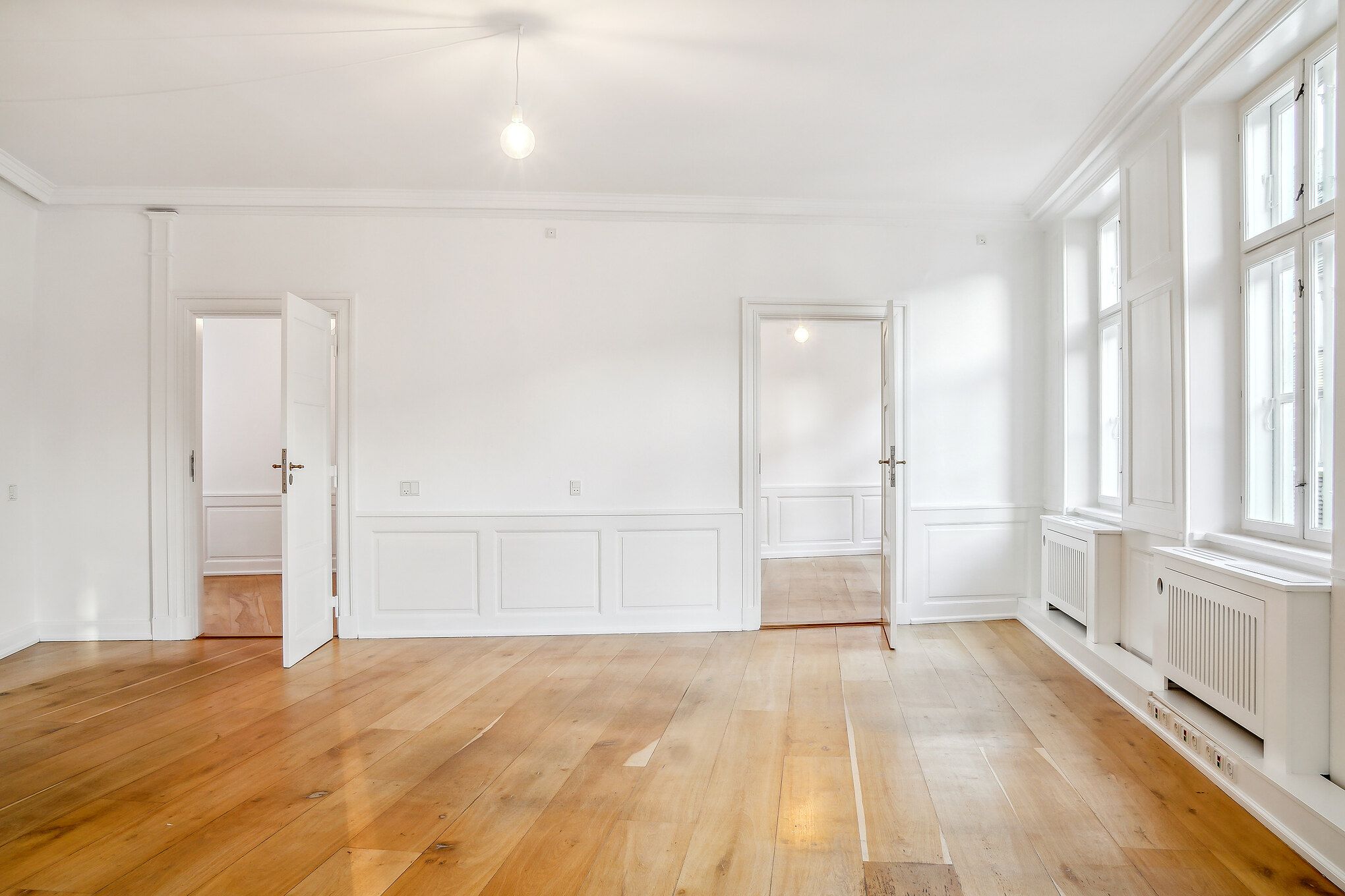 Et moderne kontor med hvide vægge og trægulve. Et lysekroner hængende fra loftet og vinduer med hvide ruder. To åbne døre fører til en lysere rum. En gulvradiator er installeret i væggen.