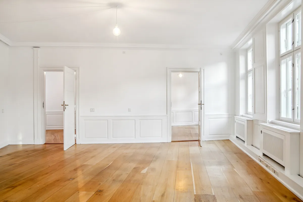 Et moderne kontor med hvide vægge og trægulve. Et lysekroner hængende fra loftet og vinduer med hvide ruder. To åbne døre fører til en lysere rum. En gulvradiator er installeret i væggen.