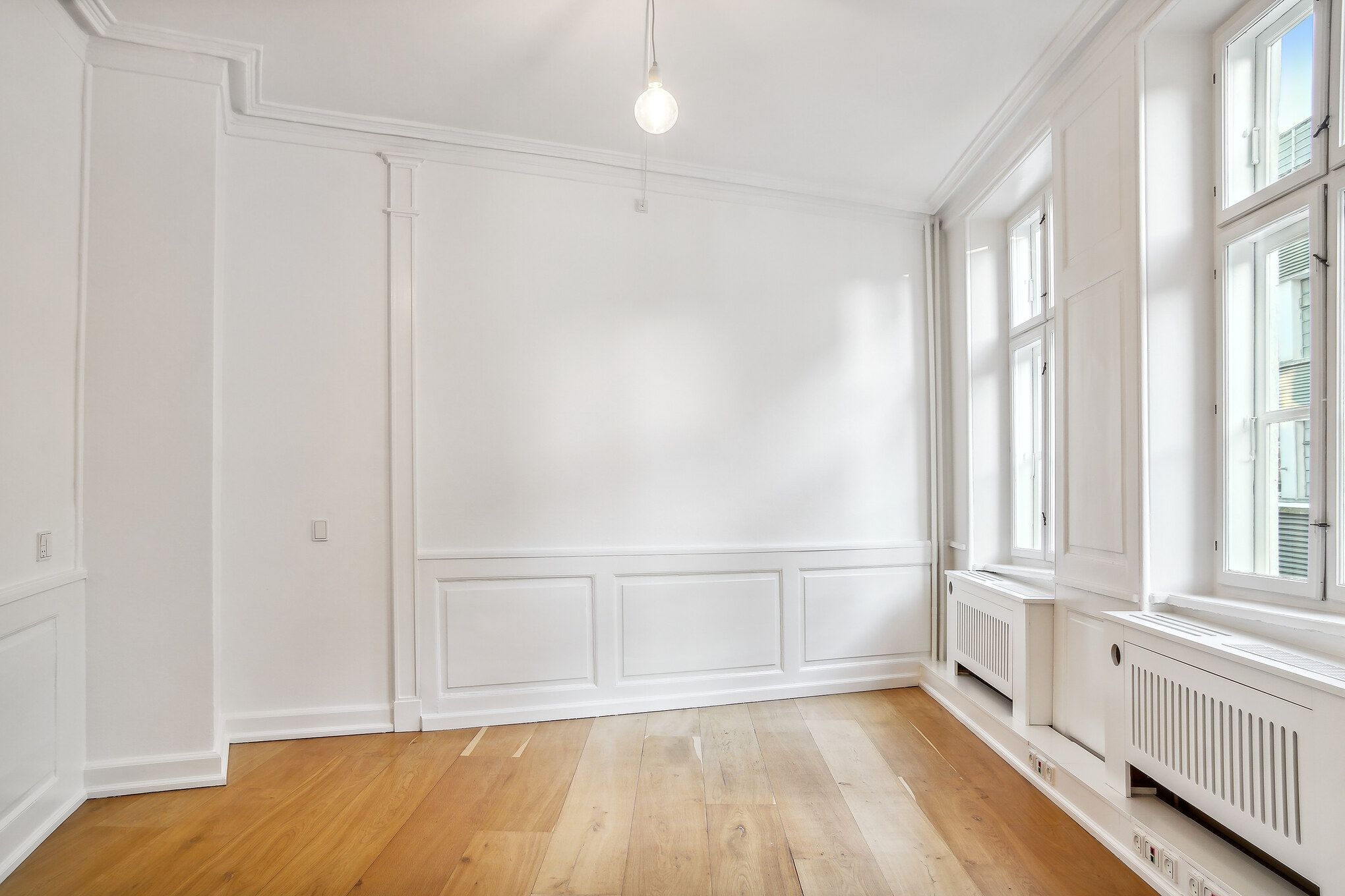 Lyst og stort kontorlokal med højloft og trægulv, hvidt panel og radiatorer, hvor lysepunkter og vinduer giver naturligt lys