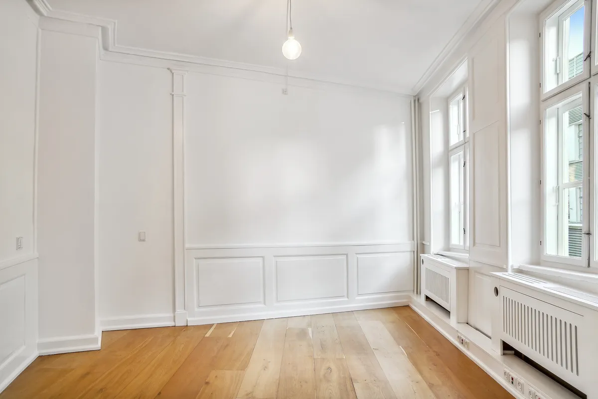 Lyst og stort kontorlokal med højloft og trægulv, hvidt panel og radiatorer, hvor lysepunkter og vinduer giver naturligt lys