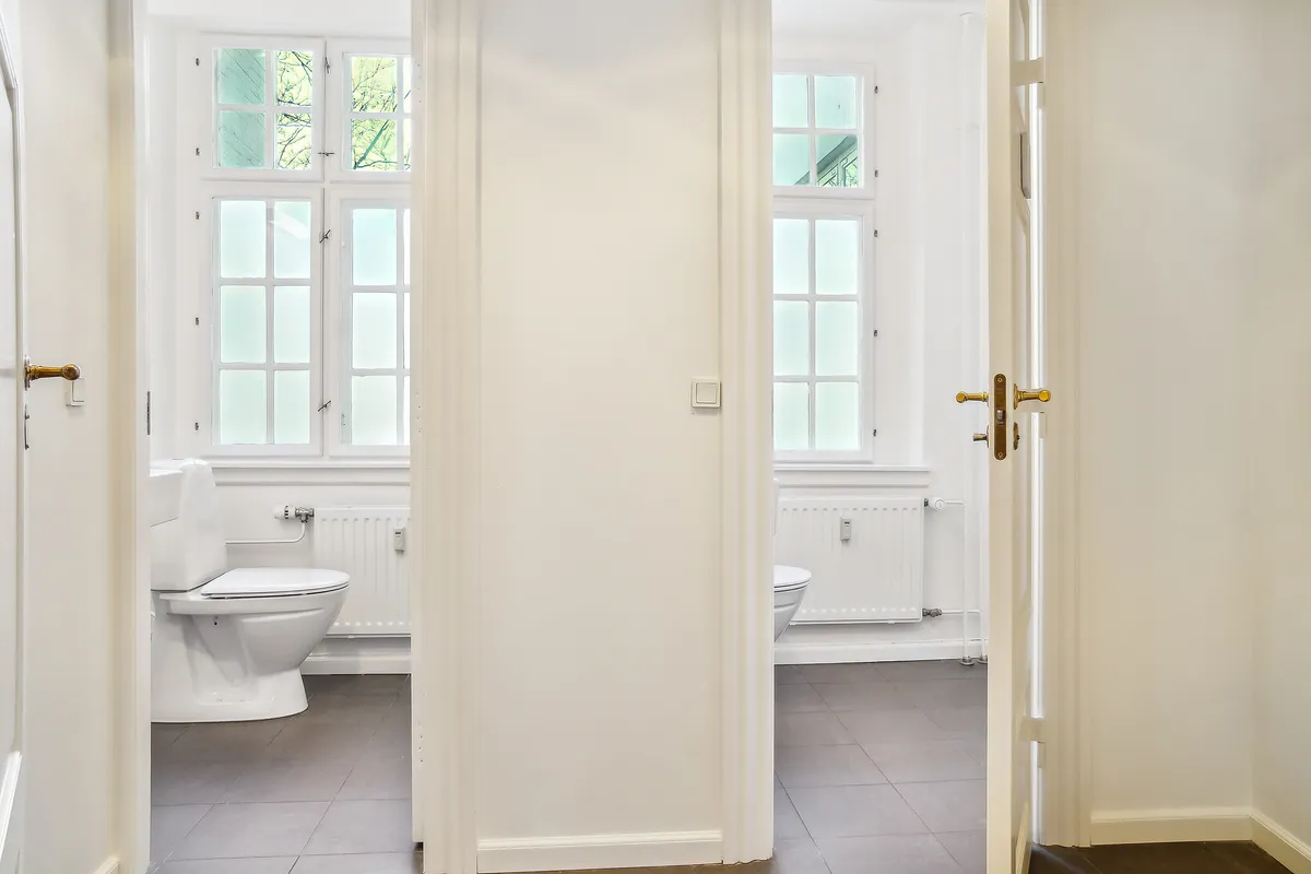 Kontor med høj gulvstand og lyse vinduer. De to toiletrum med moderne toilet og radiatorer. Gulvtæppe og lyse lofter. Elegante guldfarvede døre med lukkelås.