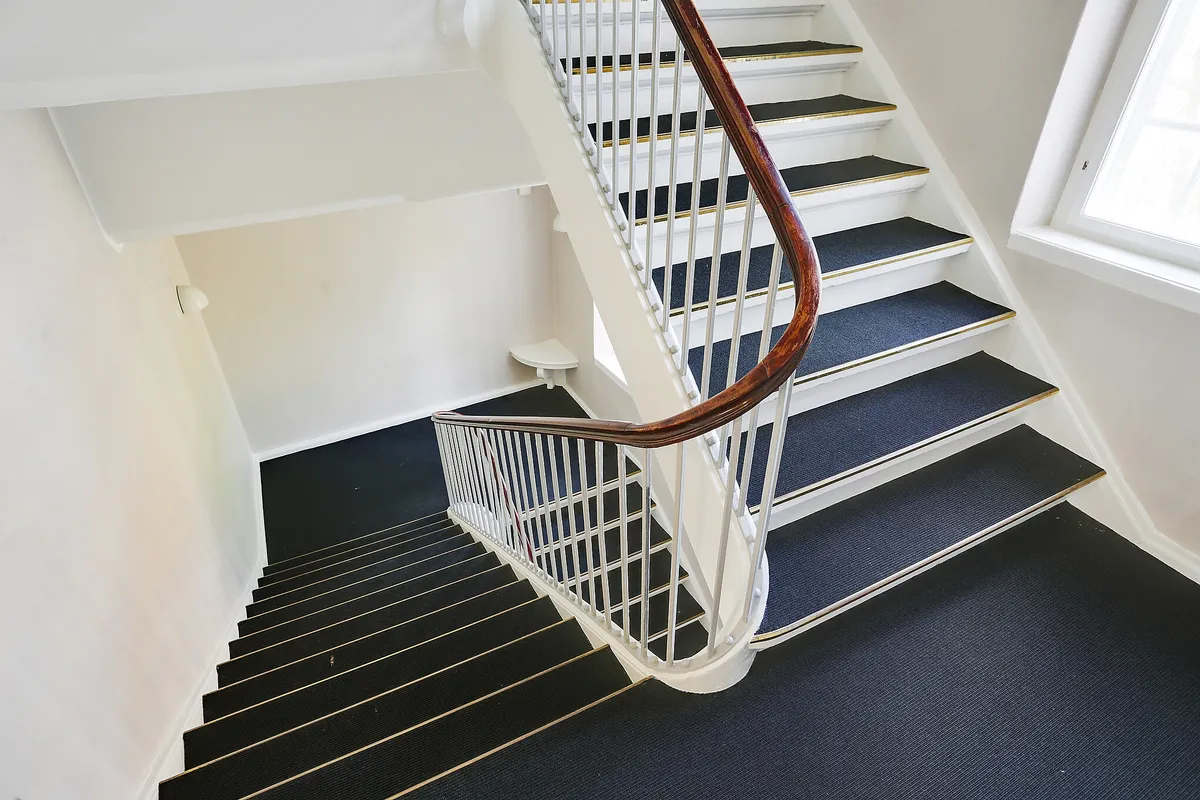 Moderne kontorsteg, hvor hvide vægge og blå gulv mødes. Støjlette trapper med metalgevær og træhåndtag. Et enkelt hvidt toilet på siden og et vindue til højre.