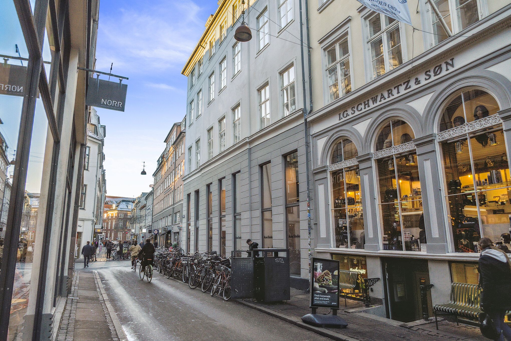Mennesker går og cykler ad en gågade. Butikker med store ruder. En række cykler står parkeret. Lysskiltene "NORR" og "G. SCHWARTZ & SON"